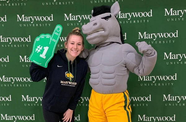 A female student poses with a Pacer mascot with a Marywood University backdrop in the background.